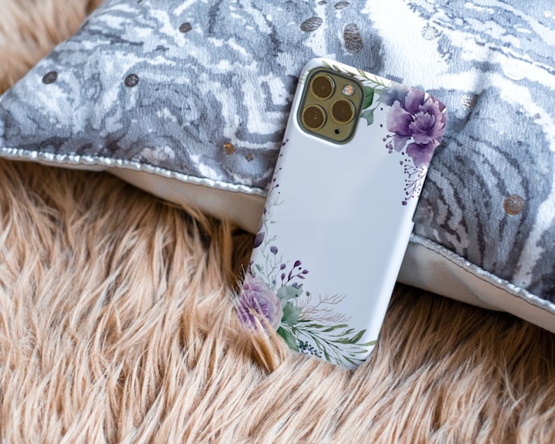 May include: A white phone case with a floral design featuring purple flowers and green leaves. The case has a camera lens cutout and is lying on a brown fuzzy surface.