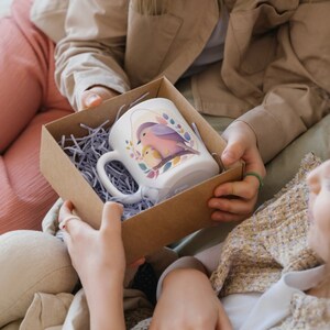 May include: A white ceramic mug with a colourful bird design is being presented as a gift in a brown cardboard box with shredded paper. The mug has a handle and a pink bird with a yellow chick on a branch.