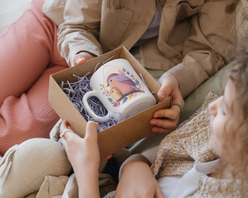 May include: A white ceramic mug with a colourful bird design is being presented as a gift in a brown cardboard box with shredded paper. The mug has a handle and a pink bird with a yellow chick on a branch.