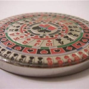 May include: A silver-colored circular casino chip with a red, black, and green design. The chip features a roulette wheel with numbers and playing card suits.