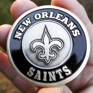 May include: A silver and black coin with the words "NEW ORLEANS SAINTS" around the edge. A fleur-de-lis is in the center.