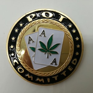 May include: A gold-colored round coin with black lettering around the edge that reads "POT COMMITTED". The center of the coin features two playing cards with the ace of spades on each card. A green marijuana leaf is on the card on the right.