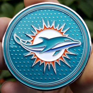 May include: A silver coin with a teal blue background and a white dolphin jumping out of a bright orange sun. The dolphin has a blue fin and tail.