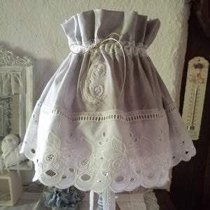 Peut inclure: Un abat-jour blanc avec un dessus en tissu gris et une bordure en dentelle blanche. L'abat-jour est noué avec un ruban de ficelle brun. La base de la lampe est grise et a une base carrée.