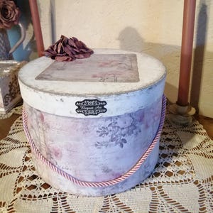 May include: A round, vintage-style hat box with a floral design in shades of pink and purple. The box has a white lid and a pink rope handle. The text "Elegant Art" is visible on the front.