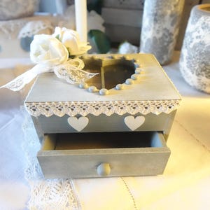 Puede incluir: Una caja de madera gris con un recorte en forma de corazón y un cajón. La caja está decorada con encaje blanco y tiene corazones blancos en la parte delantera. Un ramo de flores blancas está encima de la caja.
