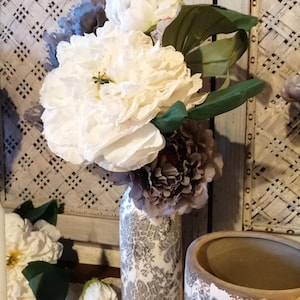 Puede incluir: Un ramo de flores artificiales blancas y grises en un jarrón de cerámica decorativo con un patrón floral. El jarrón tiene un acabado blanco desgastado con detalles florales grises.