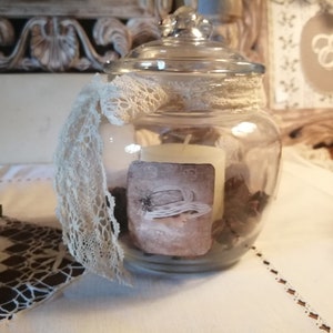 Pode incluir: Um frasco de vidro transparente com tampa, decorado com uma fita de renda branca e uma fita bege. Dentro do frasco há uma vela branca e elementos decorativos marrons.