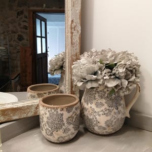 Puede incluir: Una jarra de cerámica blanca con un patrón floral y un arreglo de flores artificiales grises. La jarra tiene un asa y un pico. La jarra está sentada sobre una superficie blanca.