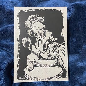 May include: Black and white illustration of a Muppet character, possibly a Fraggle Rock character, on a light gray card. The character is seated and holding a small object. The artist's name, "SCALES", is printed in the lower left corner.