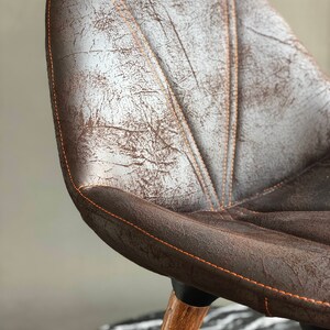 May include: Close-up of a brown leather chair with orange stitching. The chair has a distressed finish and a wooden leg. The seat has a quilted design. The chair is a piece of furniture.