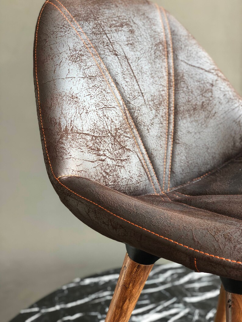 May include: Close-up of a brown leather chair with orange stitching. The chair has a distressed finish and a wooden leg. The seat has a quilted design. The chair is a piece of furniture.