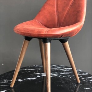 May include: A modern chair with a rust-colored, stitched seat and backrest. The chair has three wooden legs and a black base. The chair is designed with a curved back for comfort and support. The chair is suitable for a dining room or living room.