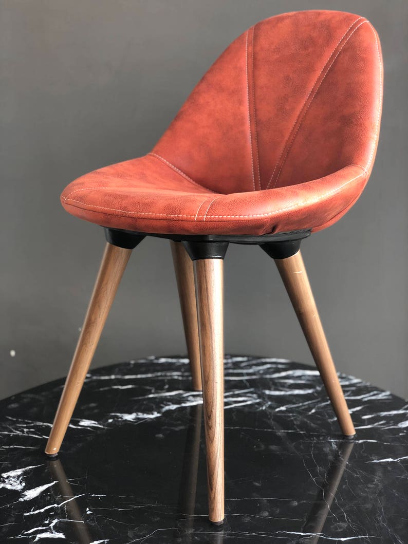 May include: A modern chair with a rust-colored, stitched seat and backrest. The chair has three wooden legs and a black base. The chair is designed with a curved back for comfort and support. The chair is suitable for a dining room or living room.