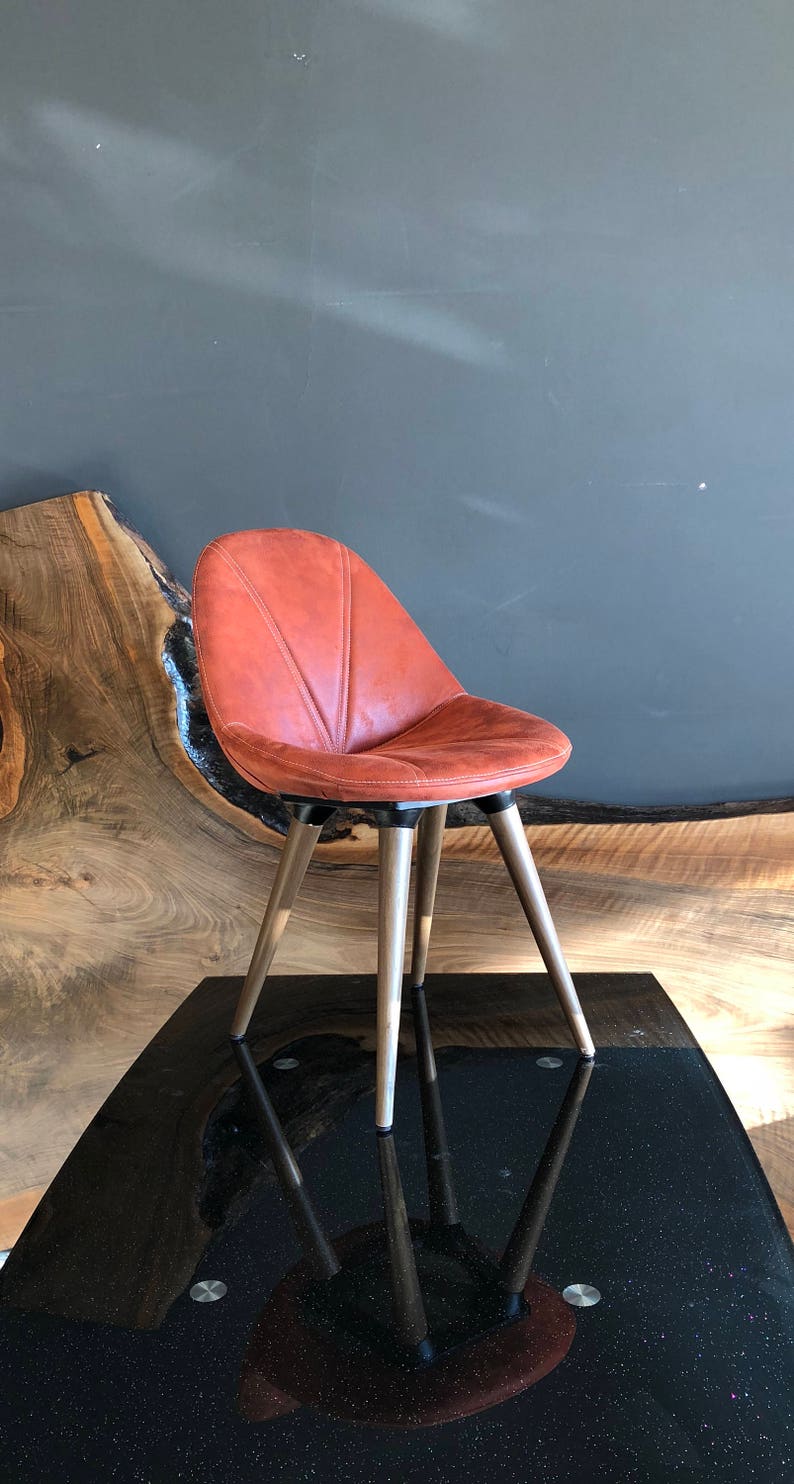 May include: An orange-brown leather chair with a rounded back and wooden legs. The chair is set on a black table with a reflective surface. The background features a dark gray wall and a wooden slab.