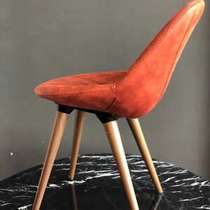 May include: An orange-brown upholstered chair with a curved back and seat. The chair has four wooden legs and is set on a black and white marble-patterned surface. The chair's design is modern and minimalist.