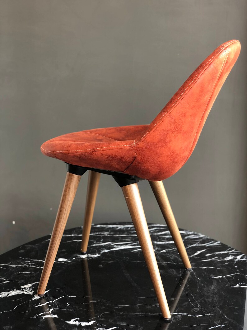 May include: An orange-brown upholstered chair with a curved back and seat. The chair has four wooden legs and is set on a black and white marble-patterned surface. The chair's design is modern and minimalist.