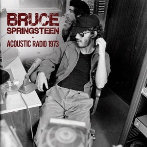 May include: Black and white photograph of Bruce Springsteen, wearing a black jacket and sunglasses, seated in a chair with a microphone in front of him. The text "Bruce Springsteen Acoustic Radio 1973" is visible at the top of the image.
