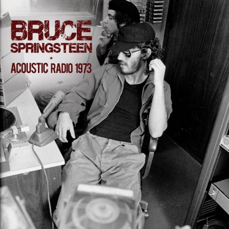 May include: Black and white photograph of Bruce Springsteen, wearing a black jacket and sunglasses, seated in a chair with a microphone in front of him. The text "Bruce Springsteen Acoustic Radio 1973" is visible at the top of the image.