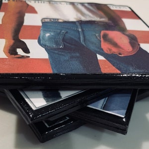 May include: A stack of four coasters with a black border and a printed image of a man wearing blue jeans and a white shirt. The image is of the album cover for "Born in the U.S.A." by Bruce Springsteen.