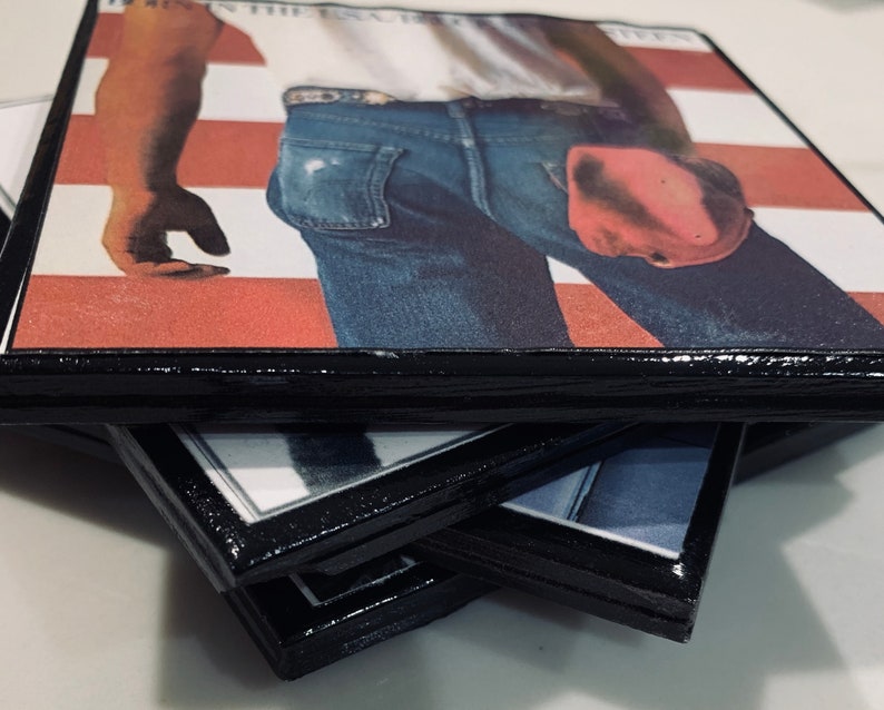 May include: A stack of four coasters with a black border and a printed image of a man wearing blue jeans and a white shirt. The image is of the album cover for "Born in the U.S.A." by Bruce Springsteen.
