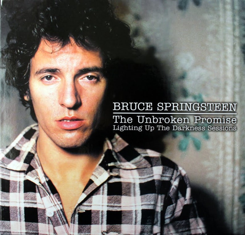 May include: A black and white checked shirt worn by Bruce Springsteen, with the text "BRUCE SPRINGSTEEN The Unbroken Promise Lighting Up The Darkness Sessions" on a black background.
