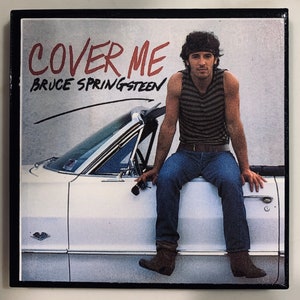 May include: A vintage album cover featuring a man in a striped tank top and jeans sitting on the bonnet of a white convertible car. The album title "COVER ME" is written in red lettering with the artist's name "BRUCE SPRINGSTEEN" written below in black lettering.