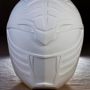 Può includere: Un casco bianco scolpito con un design dettagliato. Il casco presenta una visiera prominente, protezioni per le guance e una cresta centrale, il tutto con linee e forme intricate. Il design generale suggerisce un tema futuristico o da supereroe.