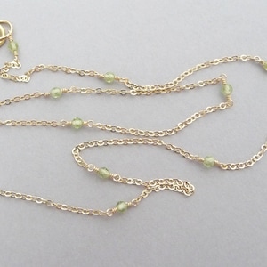 May include: A gold chain necklace with small green gemstone beads. The necklace is layered and has a lobster clasp closure.