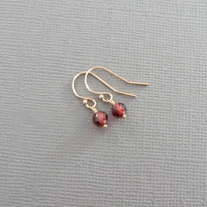 May include: A pair of gold-colored earrings with small, round, deep red gemstones. The earrings have a simple hook design, and the gemstones are securely attached. The jewelry is displayed against a neutral gray background.