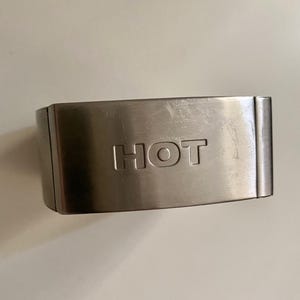 Könnte beinhalten: Ein rechteckiges Armband aus gebürstetem, silberfarbenem Metall mit dem eingeprägten Wort HOT in Großbuchstaben in der Mitte. Das Armband hat eine glatte, metallische Oberfläche mit feinen Kratzern und abgerundeten Kanten an beiden Seiten.