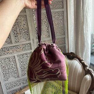 Può includere: Una pochette color prugna con chiusura a coulisse, decorata con ricami floreali dorati e lunghe frange di perline verde lime brillante che pendono dal fondo. La borsa è dotata di due manici in nastro abbinati color prugna.