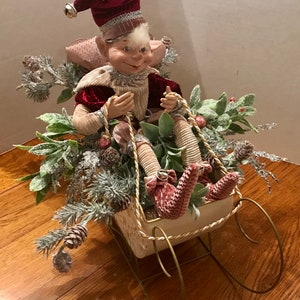 May include: A decorative Christmas elf figurine in a sleigh. The elf wears a red velvet hat with a silver trim and a matching jacket. The sleigh is cream-colored with gold accents and is filled with greenery and pinecones.