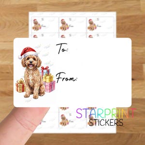 May include: A rectangular white gift label sticker featuring a watercolor illustration of a golden-colored dog wearing a red Santa hat, sitting beside a stack of colorful wrapped presents. The label includes the printed text To: and From:.