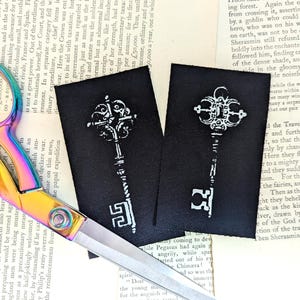 Op de afbeelding: Twee zwarte stoffen patches met witte sleutelontwerpen, naast een paar iriserende scharen op een vintage boekpagina. De sleutels hebben sierlijke details.
