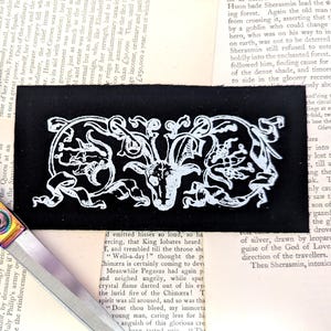 May include: A black fabric patch featuring a white, ornate design with a central goat-like figure and swirling patterns. The patch is set against a backdrop of aged book pages, with a pair of rainbow-handled scissors.