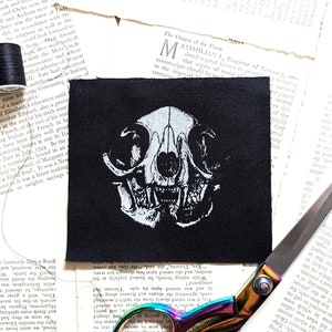 Op de afbeelding: Zwart vierkantige patch met een wit katten schedel ontwerp. Een zwarte draadspoel, schaar en pagina's met oude tekst omringen de patch. Waarschijnlijk voor naaien of knutselen.