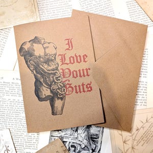 May include: A brown card with the phrase "I Love Your Guts" in red gothic script, accompanied by an anatomical drawing. It comes with a matching envelope, set on a background of old paper.