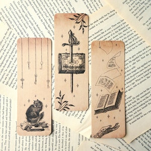 May include: Set of three vintage-style bookmarks. Each bookmark features a unique illustration: keys, a cat with books, and a sword with a crest. The bookmarks have a sepia tone and are printed on cardstock, approximately 15 cm in length.
