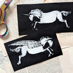 May include: Two rectangular black fabric patches featuring white screen-printed graphics. One patch displays a unicorn in a leaping pose, while the other shows a winged horse, or pegasus, in a similar dynamic stance. A pair of iridescent scissors is visible.