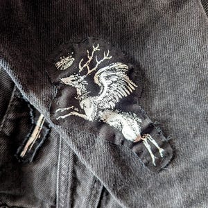 Pu&ograve; includere: Una giacca in denim grigio antracite con una toppa in tessuto cucita. La toppa presenta una grafica serigrafata bianca di un cervo alato con corna e una piccola corona sopra la testa, applicata sul tessuto scuro e testurizzato del capo.
