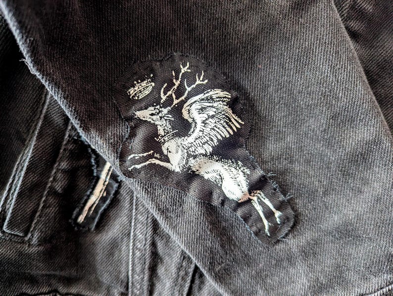 Pu&ograve; includere: Una giacca in denim grigio antracite con una toppa in tessuto cucita. La toppa presenta una grafica serigrafata bianca di un cervo alato con corna e una piccola corona sopra la testa, applicata sul tessuto scuro e testurizzato del capo.