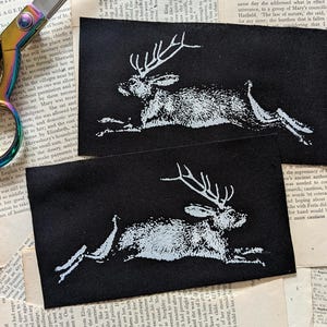 May include: Two rectangular black fabric patches featuring a white screen-printed graphic of a jackalope in a leaping motion. The patches are displayed alongside a pair of iridescent rainbow-coloured metal scissors with sharp blades.