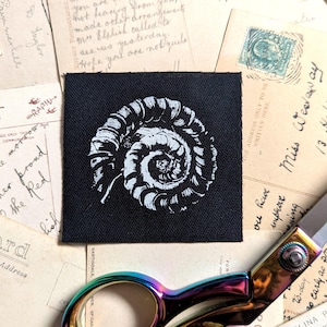 Könnte beinhalten: Ein schwarzer Aufnäher mit einer weißen, detaillierten Abbildung eines Nautilus-Gehäuses. Umgeben von Vintage-Postkarten und einer Regenbogen-Schere, erzeugt das Bild eine nostalgische Ästhetik.