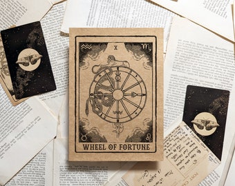 May include: A tarot card with the Wheel of Fortune design, featuring a central wheel with a snake, surrounded by zodiac symbols and clouds. The card is tan with black ink. Two other cards with a moth and moon design are also visible.