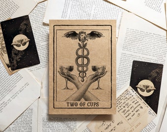 May include: A tarot card with a vintage aesthetic, featuring a central image of two hands holding chalices, with a caduceus symbol above. The card is labeled "TWO OF CUPS" and is surrounded by other cards and aged paper.