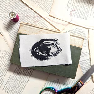 May include: A black and white eye design printed on a small piece of white fabric, placed on a green book. The image is surrounded by aged book pages, a spool of pink thread, and a pair of scissors.