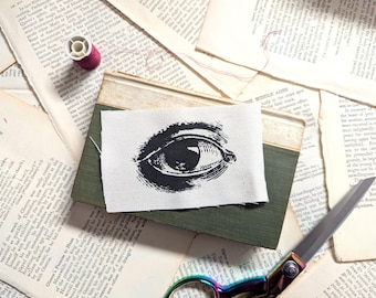 May include: A rectangular piece of off-white fabric featuring a black ink illustration of a human eye. The fabric patch is placed on top of a green book, surrounded by scattered pages of text, a small spool of magenta thread, and metal scissors.