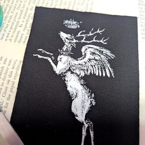 Pu&ograve; includere: Una toppa rettangolare in tessuto nero con un'illustrazione serigrafata bianca di un cervo alato che indossa una corona. Il cervo &egrave; raffigurato in posizione rampante con la testa rivolta verso l'alto verso una seconda corona sopra le corna.
