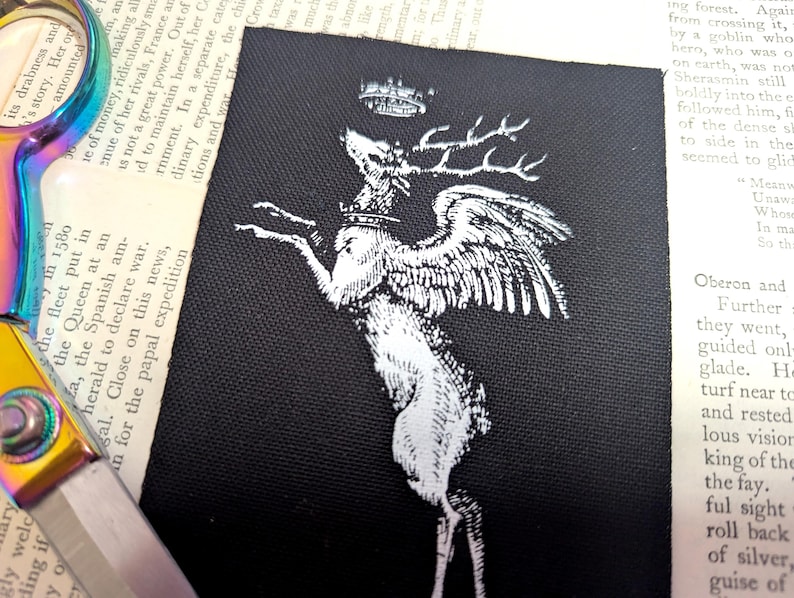 Pu&ograve; includere: Una toppa rettangolare in tessuto nero con un'illustrazione serigrafata bianca di un cervo alato che indossa una corona. Il cervo &egrave; raffigurato in posizione rampante con la testa rivolta verso l'alto verso una seconda corona sopra le corna.